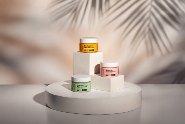 Drei farbige Tonmasken im stilvollen Studiolicht auf geometrischem Podest mit Palmenschatten im Hintergrund