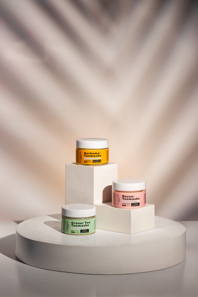 Drei bunte Tonmasken auf geometrischem Podest mit Palmenlichtspiel – moderne Produktfotografie