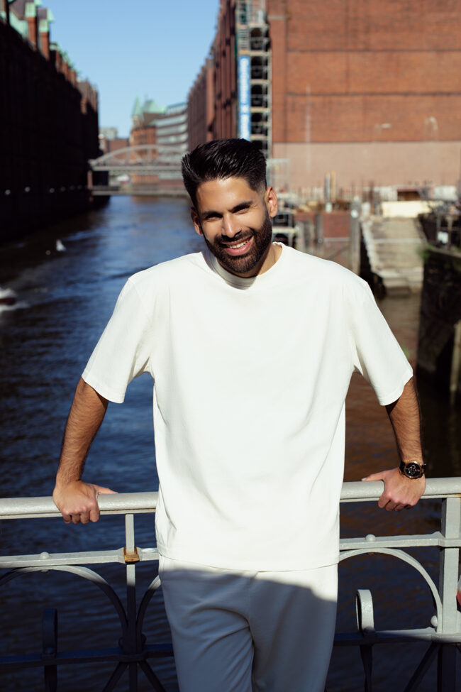 Mann in weißem Shirt steht lächelnd an Brücke in der Hamburger Speicherstadt bei Sonnenschein