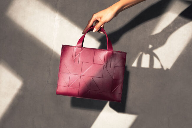 ecco-kampagne, Rote Designer-Handtasche wird von einer Hand gehalten, elegante Schatten fallen auf eine Betonoberfläche